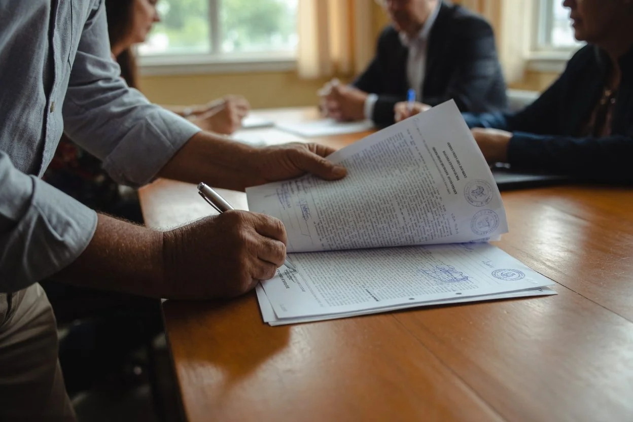 A couple's hands signing the IMM 5409 across a wooden desk, with a notary seal in the corner of the frame
